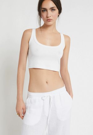 Frau, die ein weißes bauchfreies Tanktop und weiße Hose mit Kordelzug trägt, steht vor einem schlichten hellen Hintergrund.