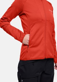 Giacca rossa con zip, in tessuto testurizzato, maniche lunghe e due tasche con zip. Abbinata a pantaloni neri con più tasche.