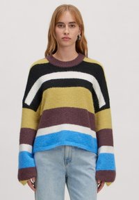 Gestreifter Pullover in Grün, Schwarz, Weiß, Braun und Blau. Hergestellt aus flauschigem Material mit Rundhalsausschnitt und oversize Ärmeln. Getragen mit hellen Jeans.