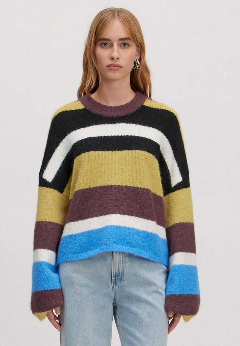 Gestreifter Pullover in Grün, Schwarz, Weiß, Braun und Blau. Hergestellt aus flauschigem Material mit Rundhalsausschnitt und oversize Ärmeln. Getragen mit hellen Jeans.