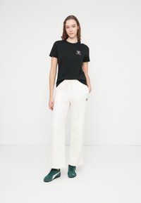 T-shirt noir à manches courtes avec petit logo, associé à un pantalon blanc à jambes larges et des chaussures de sport vertes. Tenue simple et décontractée.