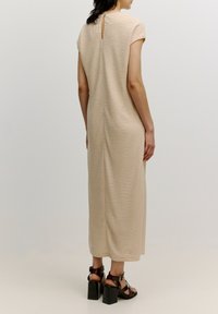 Beige, strukturiertes, knielanges Kleid mit kurzen Ärmeln, das über ein Schlüsselaugen-Detail im Rücken verfügt. Getragen mit dunklen, klobigen Sandalen mit Absatz.