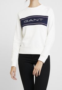 Witte katoenen sweatshirt met marineblauwe streep en "GANT" logo, ronde hals en geribbelde manchetten, gedragen over een zwarte broek.