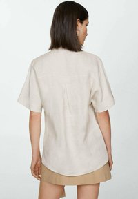 Beige linen shirt with short sleeves, rounded hemline, and back pleat detail. Smooth texture and relaxed fit.