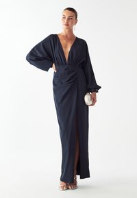 Námornícka modrá, dlhá sukňa s hlbokým V-výstrihom, stiahnutý pás, bočný rozparok a detail drapérie, skombinovaná so striebornými sandálmi.