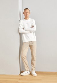 Hanorac alb cu text grafic, pantaloni bej slim-fit și adidași albi. Stând lângă un perete gri, cu brațele încrucișate, expunând un aspect casual.