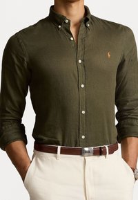 Olijfgroene blouse met lange mouwen, van zachte stof, witte knopen en een klein oranje logo. Gecombineerd met een bruine riem.