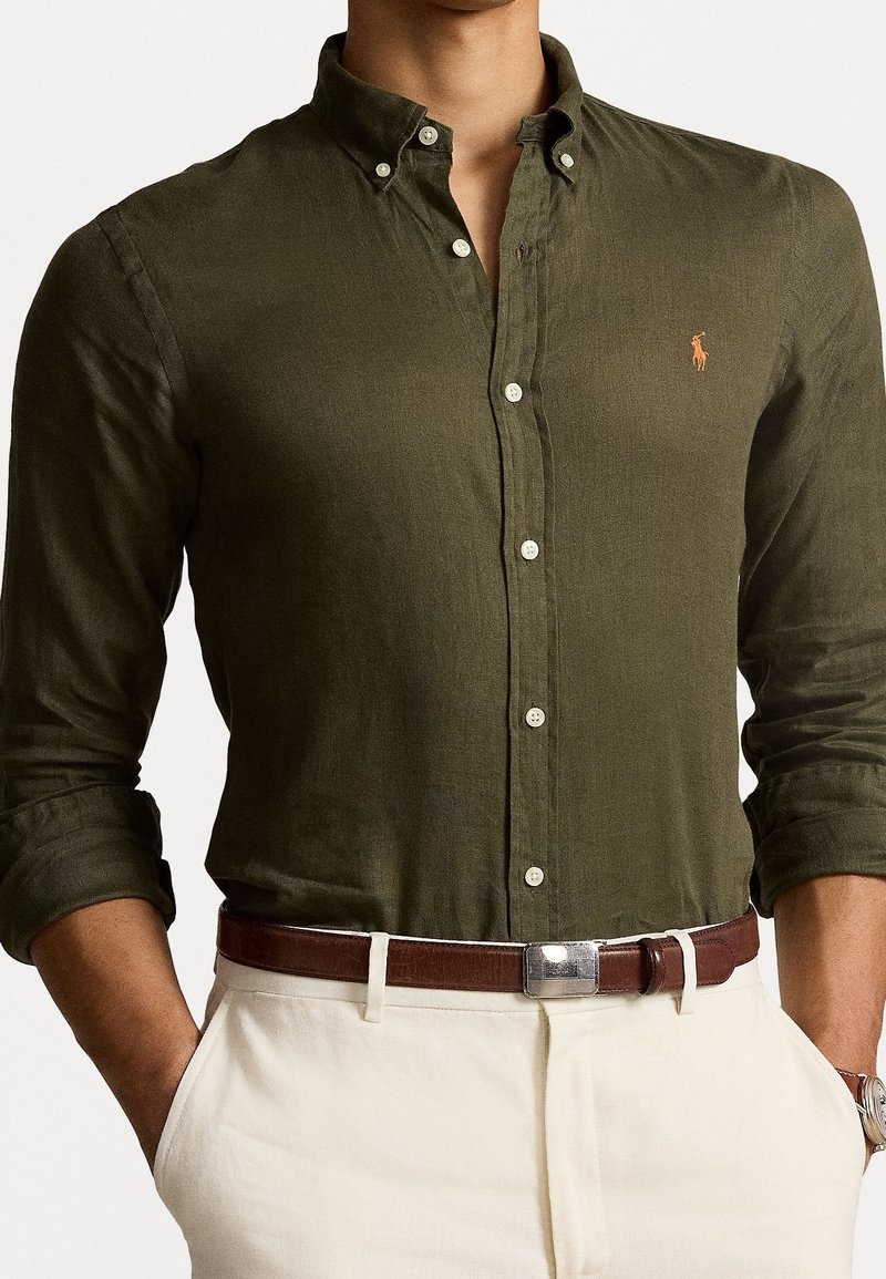 Olijfgroene blouse met lange mouwen, van zachte stof, witte knopen en een klein oranje logo. Gecombineerd met een bruine riem.