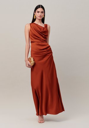 Donna con un abito in raso color ruggine che tiene una pochette dorata a forma di conchiglia, indossa orecchini dorati e tacchi aperti color nude, su uno sfondo semplice.