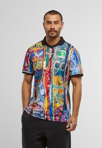 Un bărbat cu păr scurt și barbă poartă un tricou polo vibrant, cu un model multicolor, cu guler negru și pantaloni închiși, stând în fața unui fundal alb.