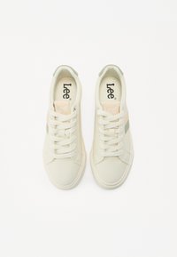 Zapatillas crema con textura suave, que presentan franjas laterales en melocotón claro y verde, puntera redonda y cordones planos. Logo visible en la lengüeta.
