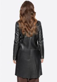 Abrigo de cuero negro con un corte ajustado, que presenta mangas largas, un corte recto y detalles de botones en los puños. Textura suave y costuras visibles.