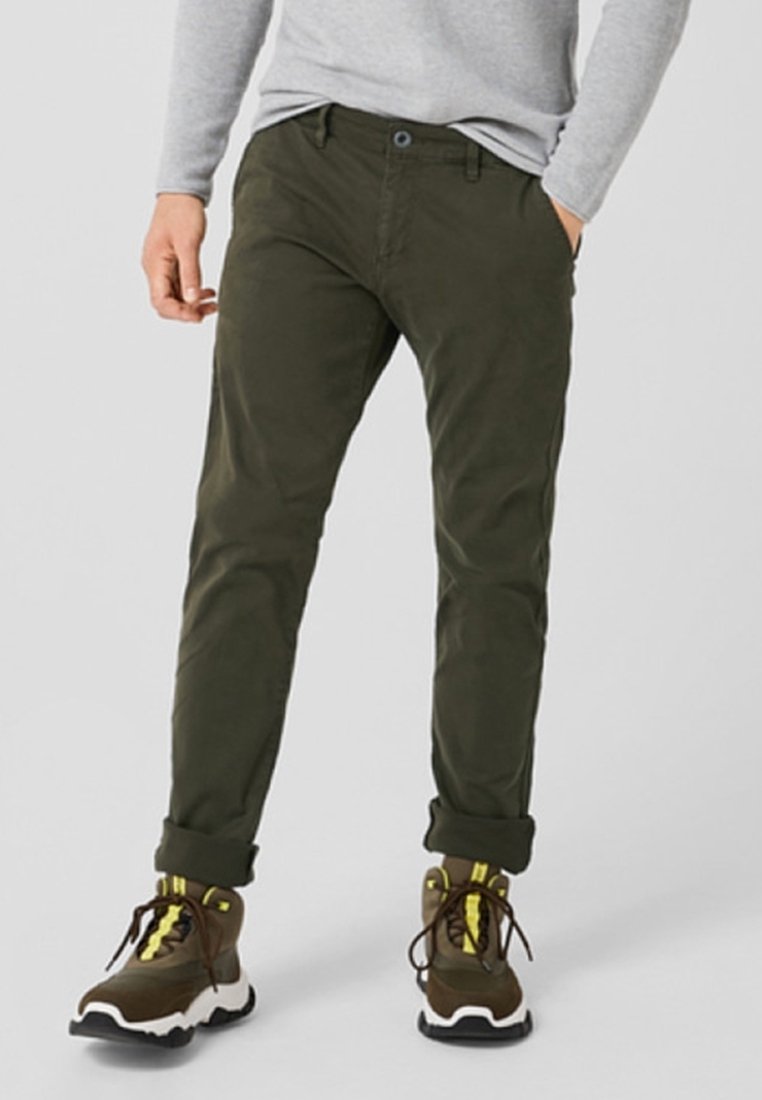 Pantalons en coton extensible vert olive avec une coupe slim, dotés d'une fermeture à bouton et de poignets retroussés, associés à des baskets marron avec des accents jaunes.