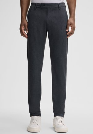 Pantaloni grigio scuro su misura con tessuto testurizzato, caratterizzati da una vestibilità slim, passanti per cintura e chiusura a bottone. Abbinati a sneakers bianche.