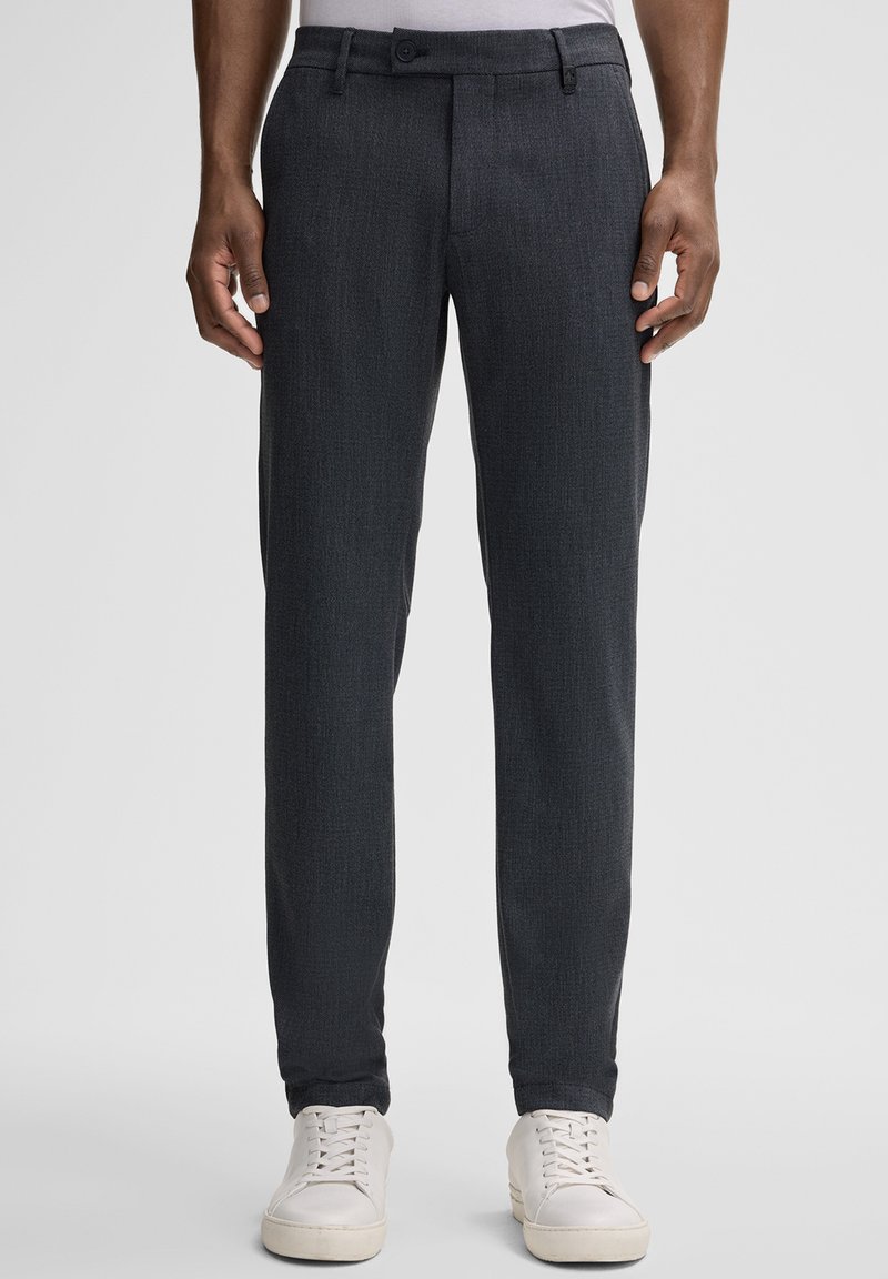 Dunkelgraue maßgeschneiderte Hose aus strukturiertem Stoff, mit schmaler Passform, Gürtelschlaufen und Knopfverschluss. Kombiniert mit weißen Sneakers.