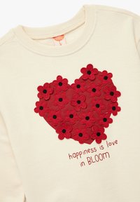 Kremowa bluza z dużym czerwonym sercem, ozdobiona czerwonymi kwiatami z czarnymi środkami, z napisem poniżej "szczęście to miłość w KWITNIENIU."