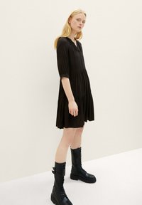 Zwarte jurk tot de knie met een knoopsluiting, driekwartmouwen en gelaagd ontwerp. Model draagt zwart veterlaarsjes.