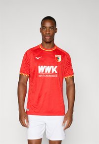 Sarkans īsām piedurknēm futbols krekls ar dzelteniem akcentiem, ar lielu baltu logotipu un vieglu teksturētu rakstu. Valkāts ar baltiem šortiem.