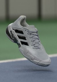 Grauer Sportschuh mit strukturiertem Finish, schwarzen Akzenten, drei schwarzen Streifen und einer gemusterten Sohle. Leicht und aerodynamisch im Design.