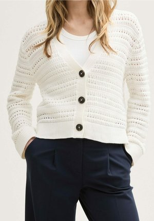 Le mannequin porte un cardigan crème en maille ajourée avec trois boutons foncés, par-dessus un haut blanc et un pantalon bleu marine.