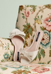 Chaussures blanches à enfiler avec un détail nœud, présentant une finition en cuir lisse et un bout ouvert, posées sur une chaise recouverte d'un tissu floral.