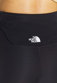 Černé sportovní legíny s hladkým povrchem. Obsahují malou zipovou kapsu a bílé logo "The North Face" na pásku.