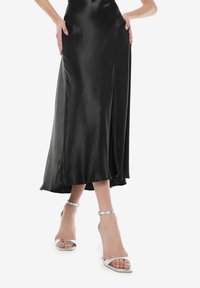 Schwarzer Satin-Midi-Rock mit glatter Textur, fließendem Design und leicht ausgestellt geschnittenem Saum. Kombiniert mit silbernen, römischen High-Heels.