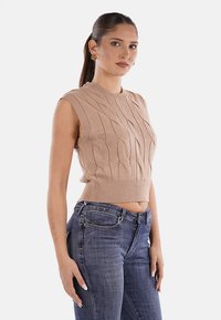 Top senza maniche beige in maglia con motivo a treccia, scollo e orlo a costina, abbinato a jeans blu a vita alta.