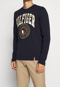 Navy pitkähihaisen t-paidan, jossa on suuri "HILFIGER"-logo kullassa, pyöreä emblem punaisella, valkoisella ja sinisellä, ribattu kaulus ja helma.