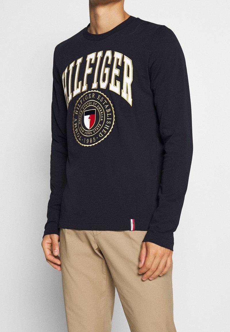 Navy pitkähihaisen t-paidan, jossa on suuri "HILFIGER"-logo kullassa, pyöreä emblem punaisella, valkoisella ja sinisellä, ribattu kaulus ja helma.