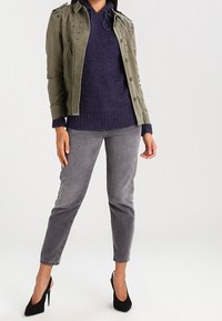 Chaqueta verde oliva con tachuelas decorativas, suéter azul marino con capucha, jeans grises y tacones altos negros. La modelo está de pie frente a un fondo neutral.