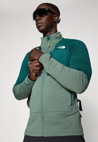 Green zip-front fleece jacket with textured fabric, contrasting teal sleeves, and black accents. Features side pockets and a high collar.