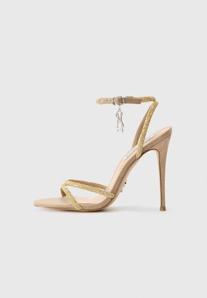 Beige high-heeled sandal with crisscross gold accents, ankle strap, and rhinestone detailing. Suede material with a pointed toe design.