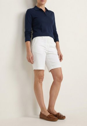 Kvinde står op ad en væg iført en mørkeblå polo med lange ærmer, hvide shorts til knæet og brune mokkasinsko.