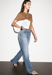 Brown cotton t-shirt, light blue flared jeans, and white textured bag. Model wearing flip-flops, walking against a neutral background.