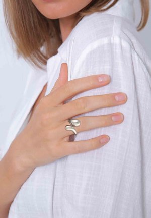 Silberner, geschwungener Ring an einer Hand, die gegen eine weiße, strukturierte Bluse ruht und ein glattes, modernes Design mit sanften Kurven zeigt.