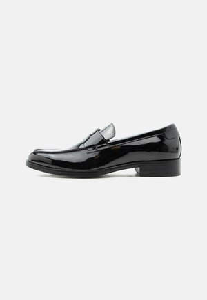 Mocassin pour homme en cuir verni noir avec talon bas, bout arrondi, et détail de bride sur le dessus, sur fond blanc.