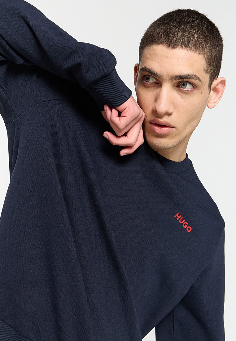 Junger Mann mit kurzen Haaren, der einen navyblauen Sweatshirt mit dem roten "HUGO"-Logo trägt und einen Arm nahe seines Gesichts gegen einen einfarbigen Hintergrund hebt.