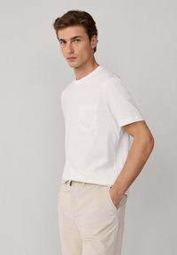 Camiseta de algodón blanca con mangas cortas, escote redondo y un bolsillo en el pecho con un sutil patrón bordado. Llevada con pantalones de color claro.