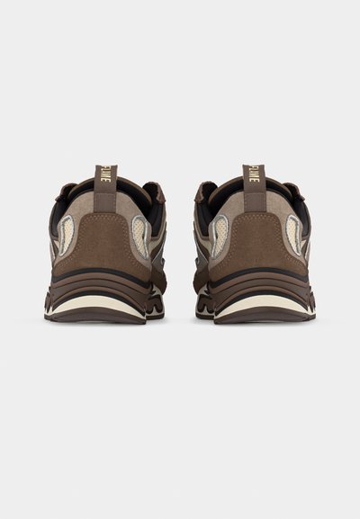 Ζευγάρι καφέ και μπεζ αθλητικών παπουτσιών που φαίνεται από πίσω, με χοντρές σόλες και θηλιές στο τακούνι, πάνω σε απλό φόντο.