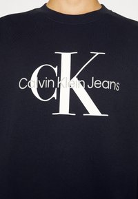Ciemnoniebieska bluza z dużymi białymi literami "CK" i mniejszym szarym napisem "Calvin Klein Jeans" na piersi.