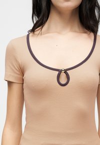 Femme portant un haut beige côtelé à manches courtes avec un col rond bordé de marron foncé et un détail de bouton doré circulaire.