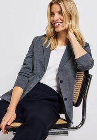 Šedý blazer s dvěma knoflíky, texturovaná tkanina, slim fit. Pod ním bílá košile doplňuje námořnické kalhoty. Sedí na tkaném křesle.