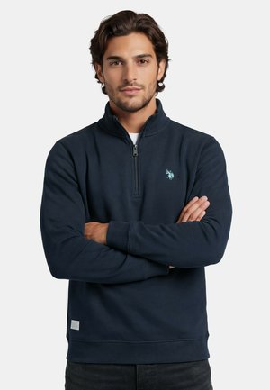 Mann mit dunklen Haaren, der einen marineblauen Pullover mit Reißverschluss und kleinem hellblauem Emblem trägt, steht mit verschränkten Armen vor einem einfarbigen Hintergrund.