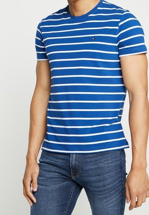 Mand iført en tætsiddende blå og hvid T-shirt med vandrette striber og mørkeblå denimjeans.