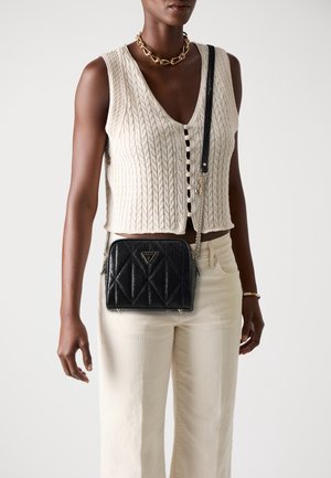 Femme portant un gilet sans manches en maille torsadée crème, un pantalon crème, un collier chaîne doré et un sac bandoulière noir matelassé avec une sangle chaîne.