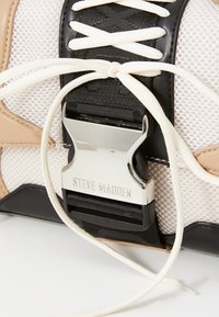 Črne in bež teniske iz mrežastega materiala, opremljene z belimi vezalkami in srebrnim kovinskim sponko s gravered "STEVE MADDEN".