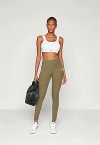 Λευκό αθλητικό crop top και ελαιόχρωμες ψηλές κολάν. Διαθέτει στενή εφαρμογή και ελαστική υφή. Μαύρη αθλητική τσάντα και λευκά αθλητικά παπούτσια.