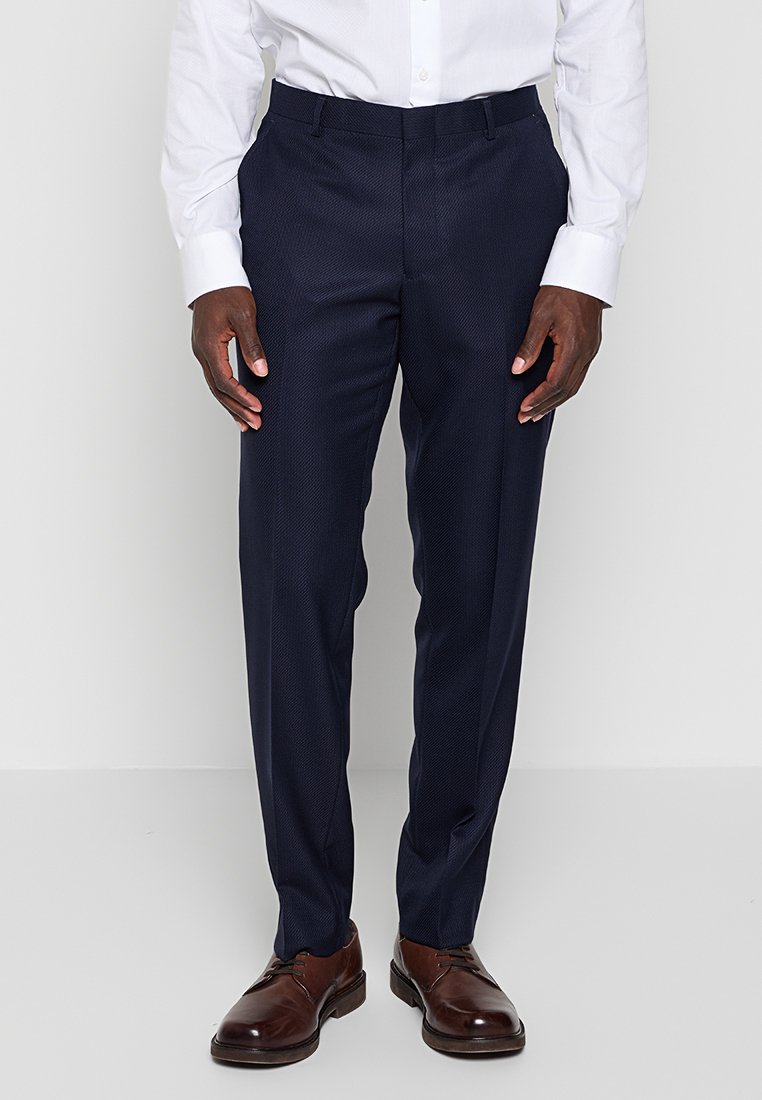 Marineblaue Anzughose, strukturiertes Gewebe, schlanker Schnitt, faltige Vorderseite, Gürtelschlaufen, kombiniert mit braunen Lederschuhen und einem weißen Hemd mit Knopfleiste.