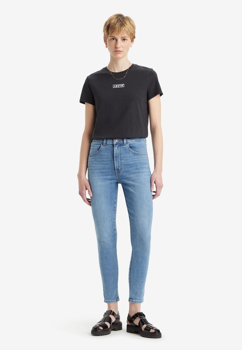 T-shirt noir en coton avec un petit logo blanc de Levi's, associé à un jean skinny taille haute bleu clair et des sandales noires à lanières.